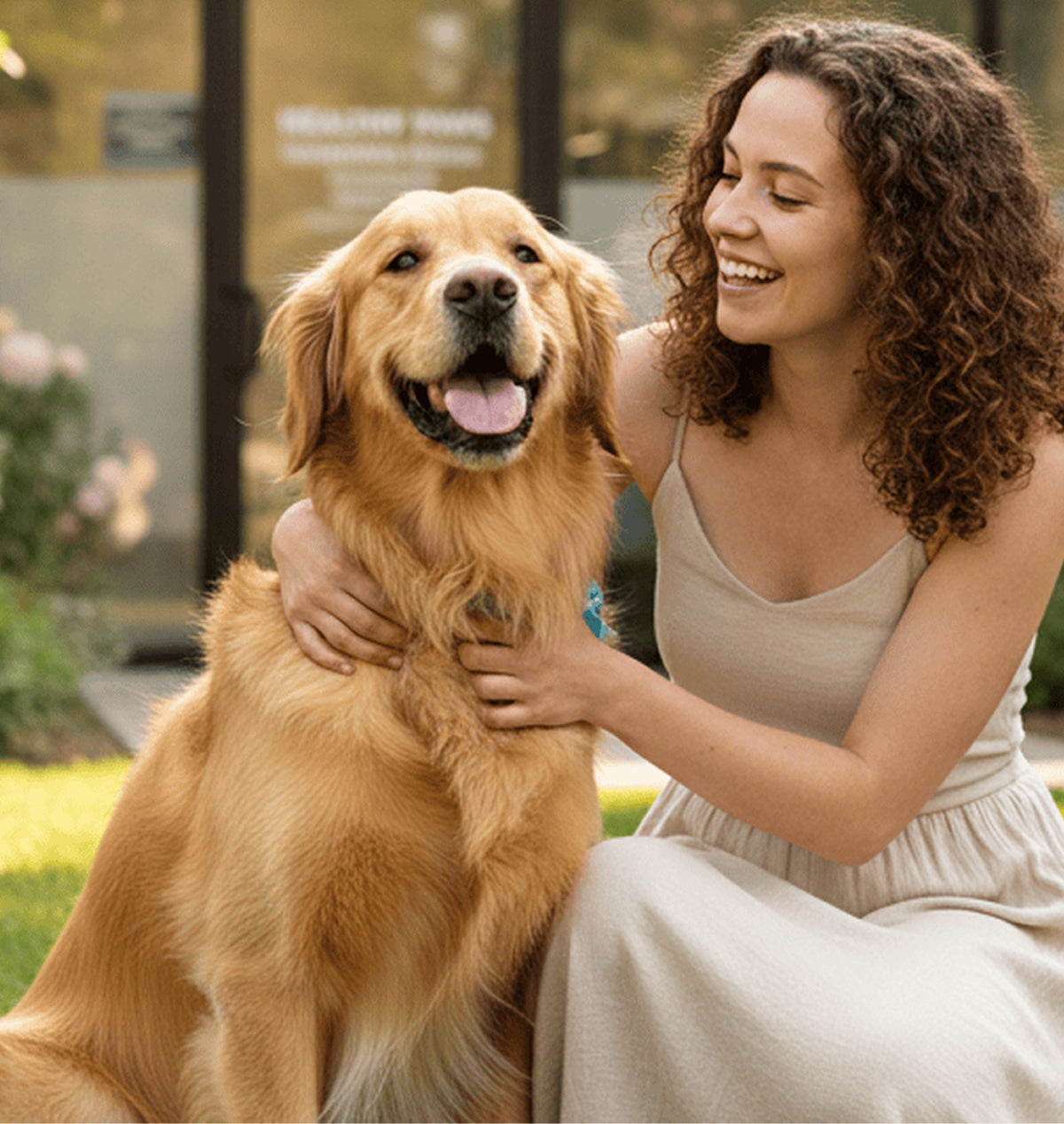 A woman in a beige sleeveless dress sits outdoors with a golden retriever dog, smiling and petting it. The background shows a softly blurred building entrance and greenery.